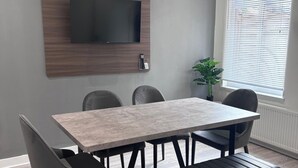 Una camera moderna con una TV a schermo piatto montata su un pannello in legno, un tavolo da pranzo con quattro sedie e una pianta in vaso vicino a una finestra con persiane.
