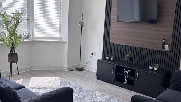 Un soggiorno moderno con televisore a schermo piatto montato su una parete rivestita in legno, un mobile TV nero con piante, una lampada da terra e divani scuri.