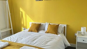 Un letto con biancheria da letto bianca e due cuscini color senape, un comodino bianco con una lampada e una parete gialla.
