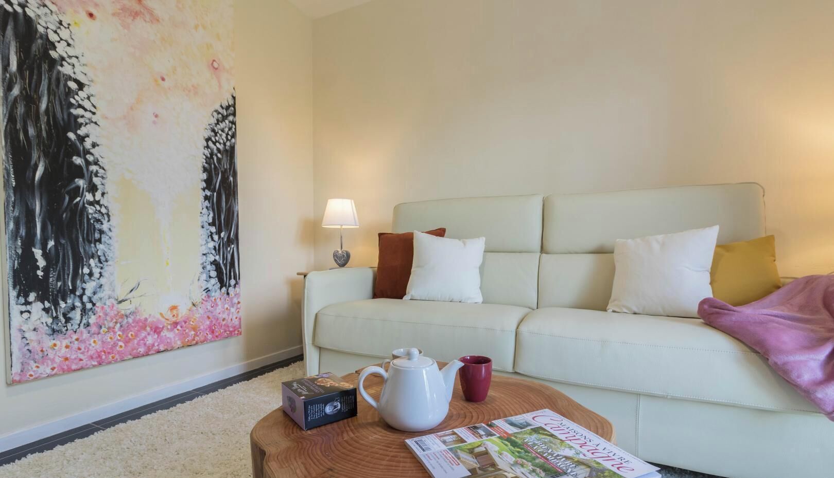 Un salon comprenant un canapé, une table basse avec une bouilloire et une tasse, une lampe et un grand tableau représentant des fleurs.