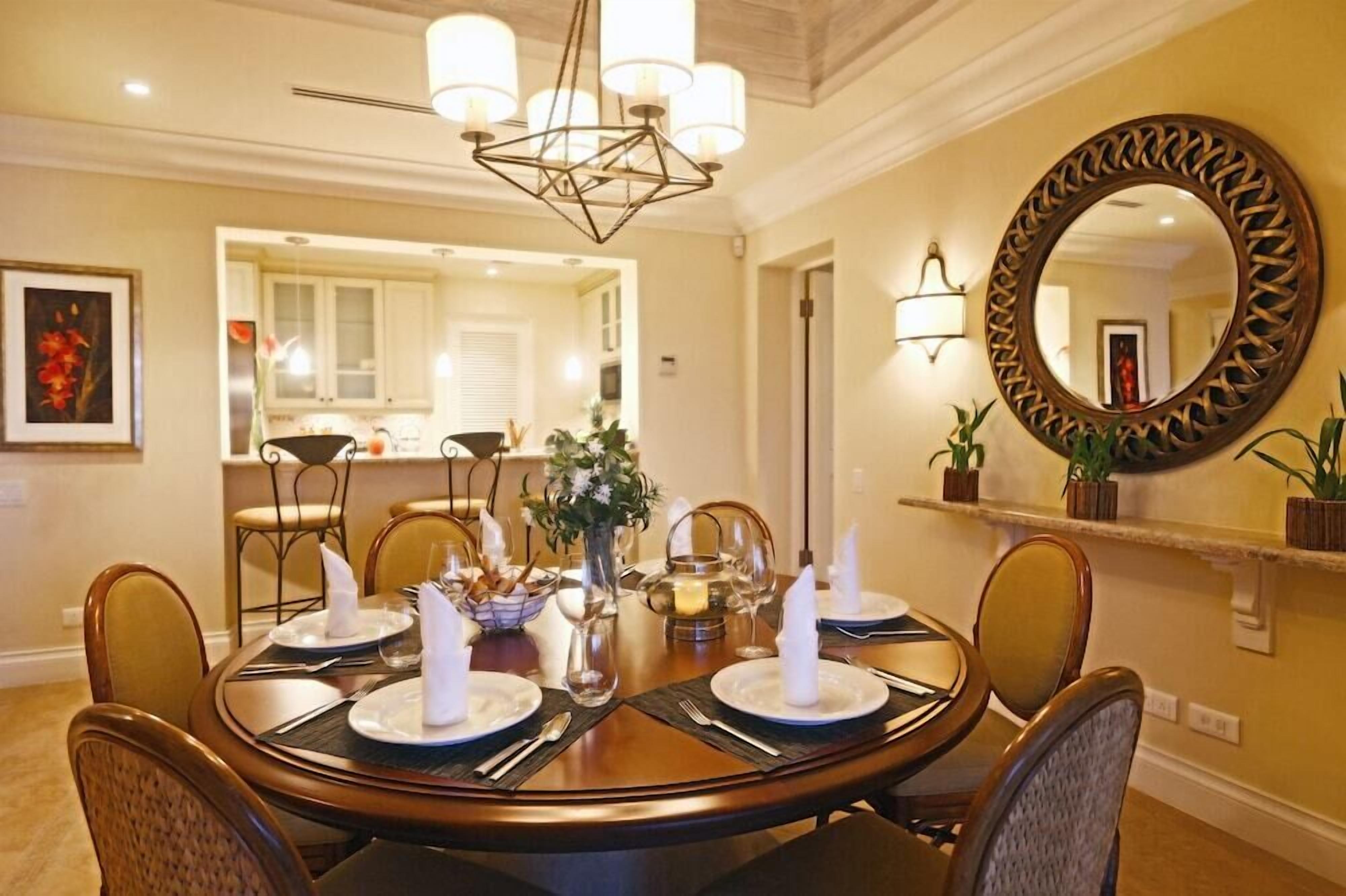 A dining area with a round table set for six, a chandelier, a mirror, and a kitchen in the background.