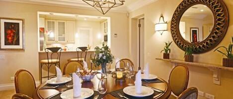 A dining area with a round table set for six, a chandelier, a mirror, and a kitchen in the background.
