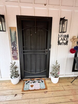 A black door with a decorative glass panel, flanked by two potted evergreen trees, a welcome sign, and a doormat with a heart design.