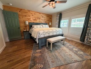 A bedroom with a wooden wall, a bed with patterned bedding, a bench, a nightstand, a ceiling fan, and a window with curtains.