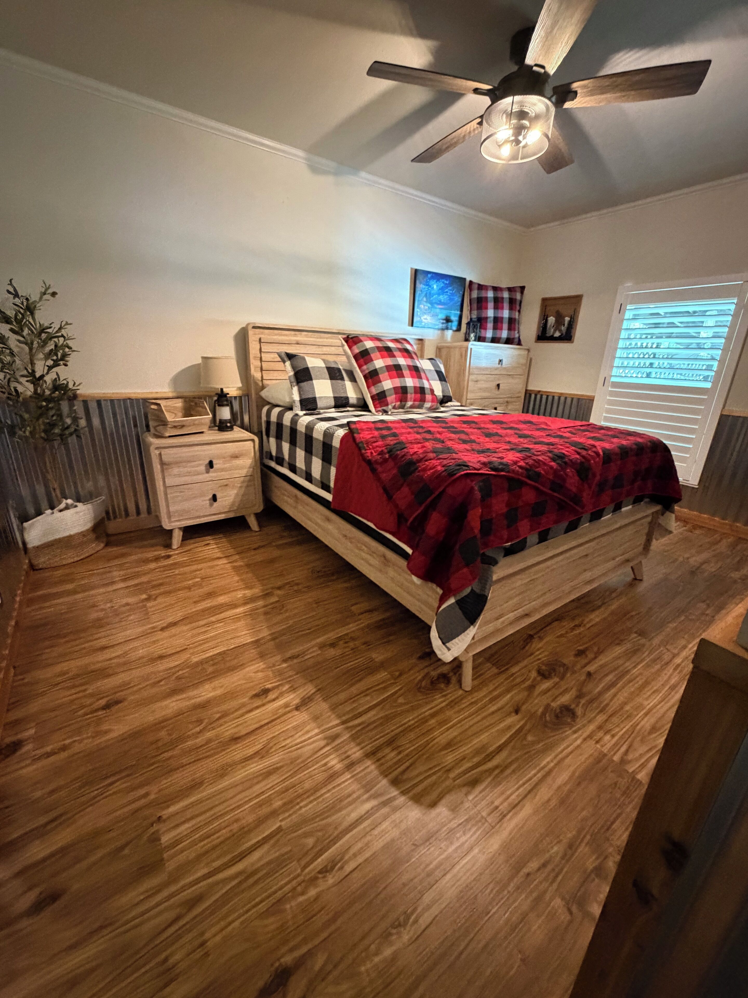 Une chambre à coucher avec un lit en bois, une table de chevet, un ventilateur de plafond et une fenêtre avec des stores.