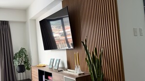 Un salón moderno con una televisión de pantalla plana montada en la pared, una mesa consola de madera con una planta en maceta y un ventanal con cortinas.
