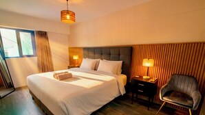 Habitación de hotel con una cama grande, mesitas de noche, una silla, un ventanal con cortinas y suelo de madera.