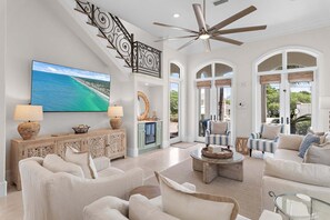 A spacious living room with a large TV, comfortable seating, a ceiling fan, and a view through the arched windows. - Blue Mountain Bella | Pool + Golf Cart + Steps to Beach (Santa Rosa Beach)