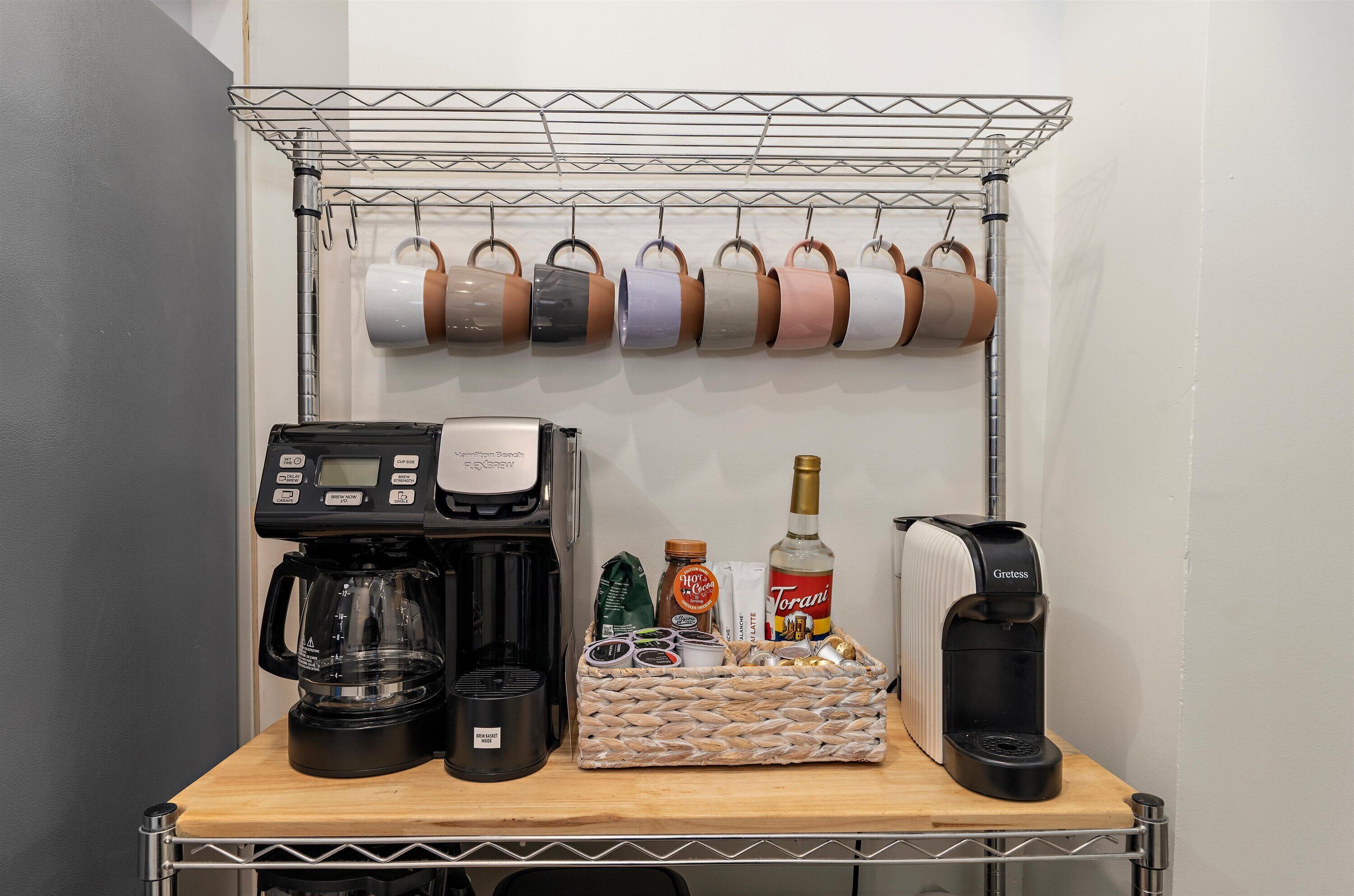 A coffee station with a coffee maker, a basket of coffee pods, a bottle of Torani syrup, and a kettle.