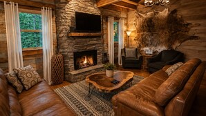 Ein gemütliches Blockhaus-Interieur mit Kamin aus Stein, Ledersofas und einer Steinwand mit einem angebrachten Fernseher.
