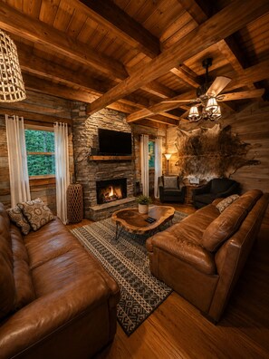 A cozy log cabin interior with a stone fireplace, leather sofas, and a stone wall with a mounted TV. - Beautiful & Perfectly Located Cabin w/ Hot Tub + Bunk Room + Game Room (Gatlinburg)
