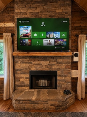 Un salon avec une cheminée en pierre, une console Xbox fixée au mur et un écran présentant une sélection de jeux.