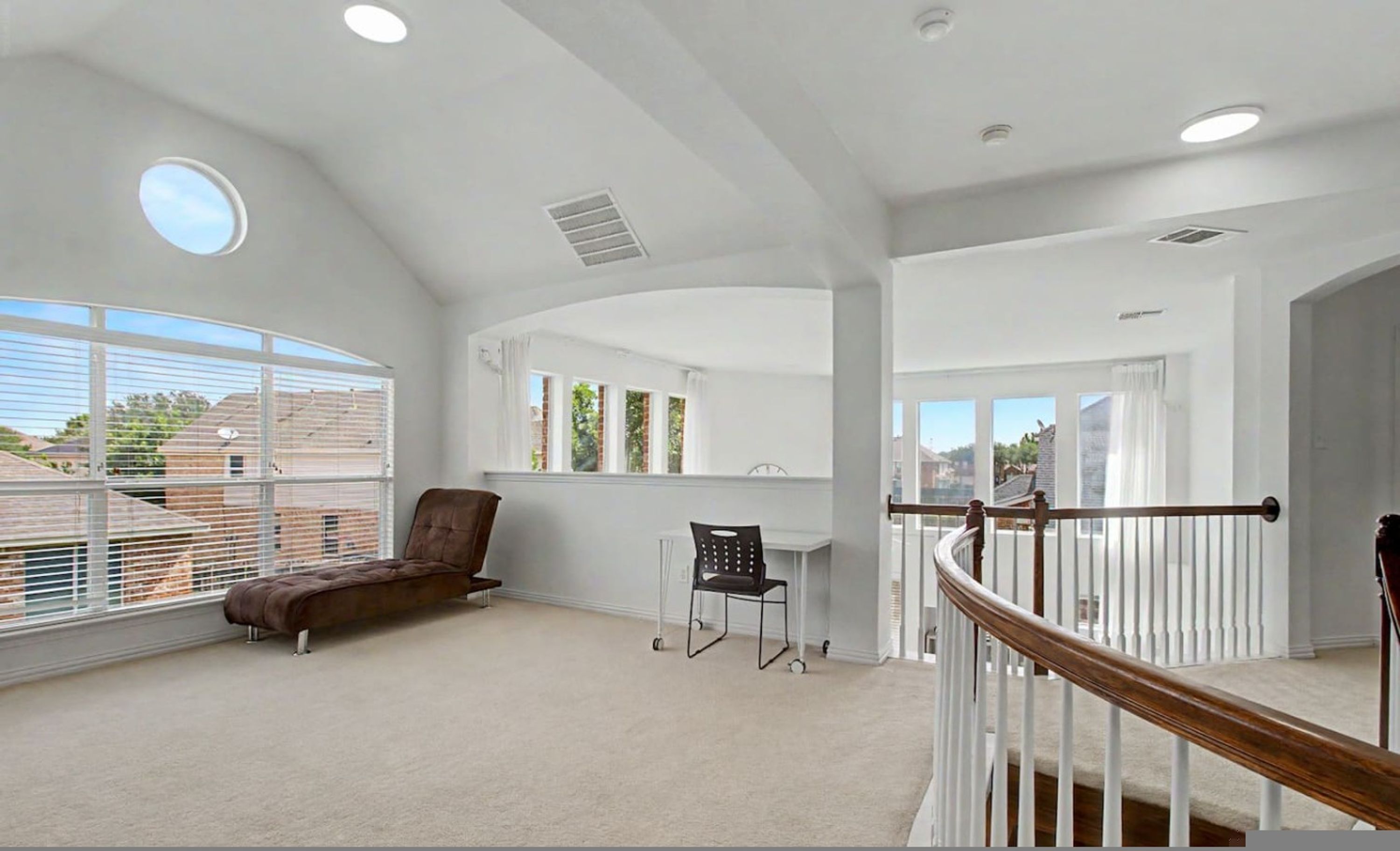 A spacious room with a high ceiling, large windows, a brown leather sofa, a small table with two chairs, and a wooden railing.