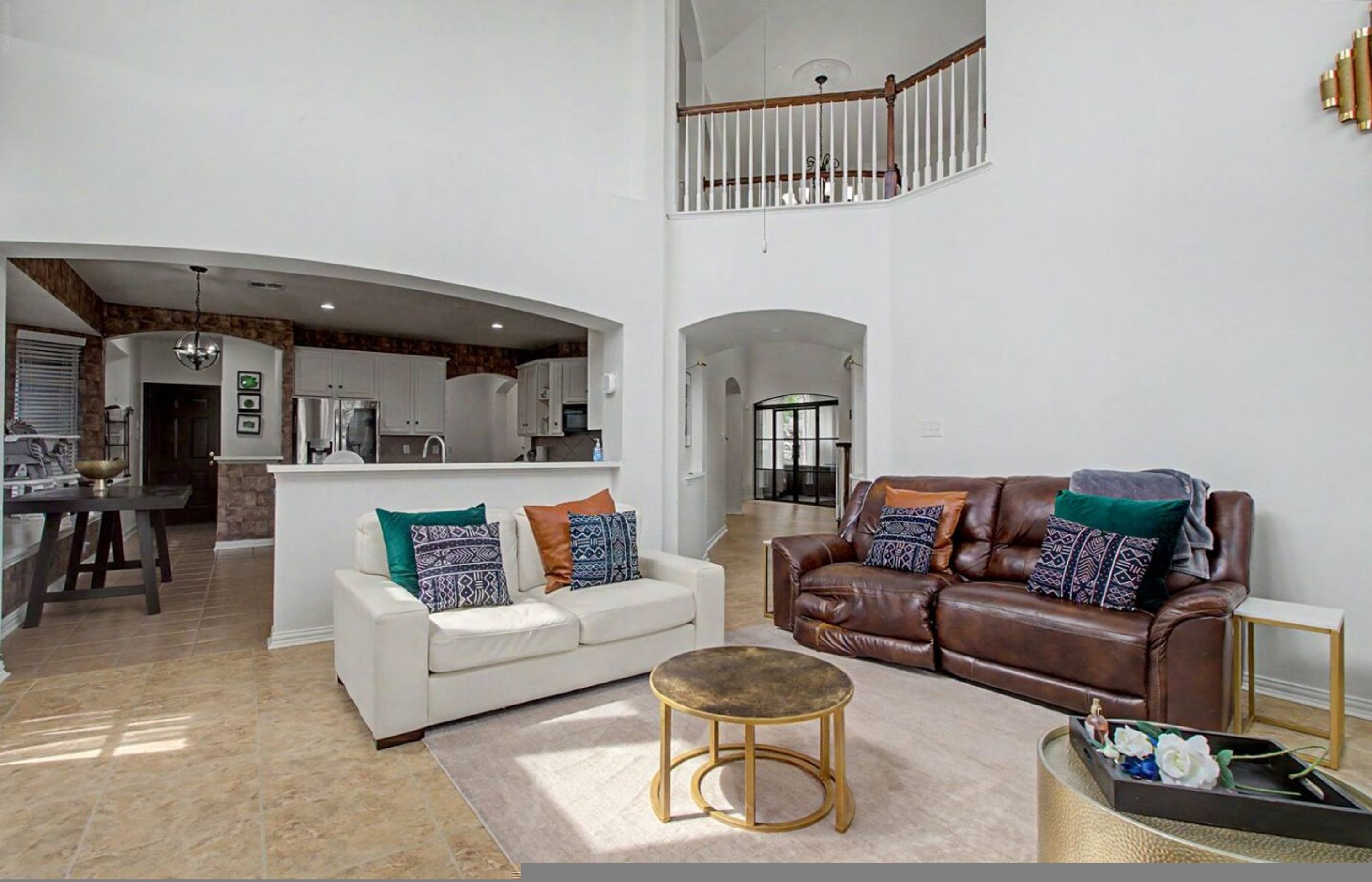 A spacious living area with a white sofa, a brown leather sofa, and a round coffee table. A kitchen is visible in the background with white cabinets and a dining area.