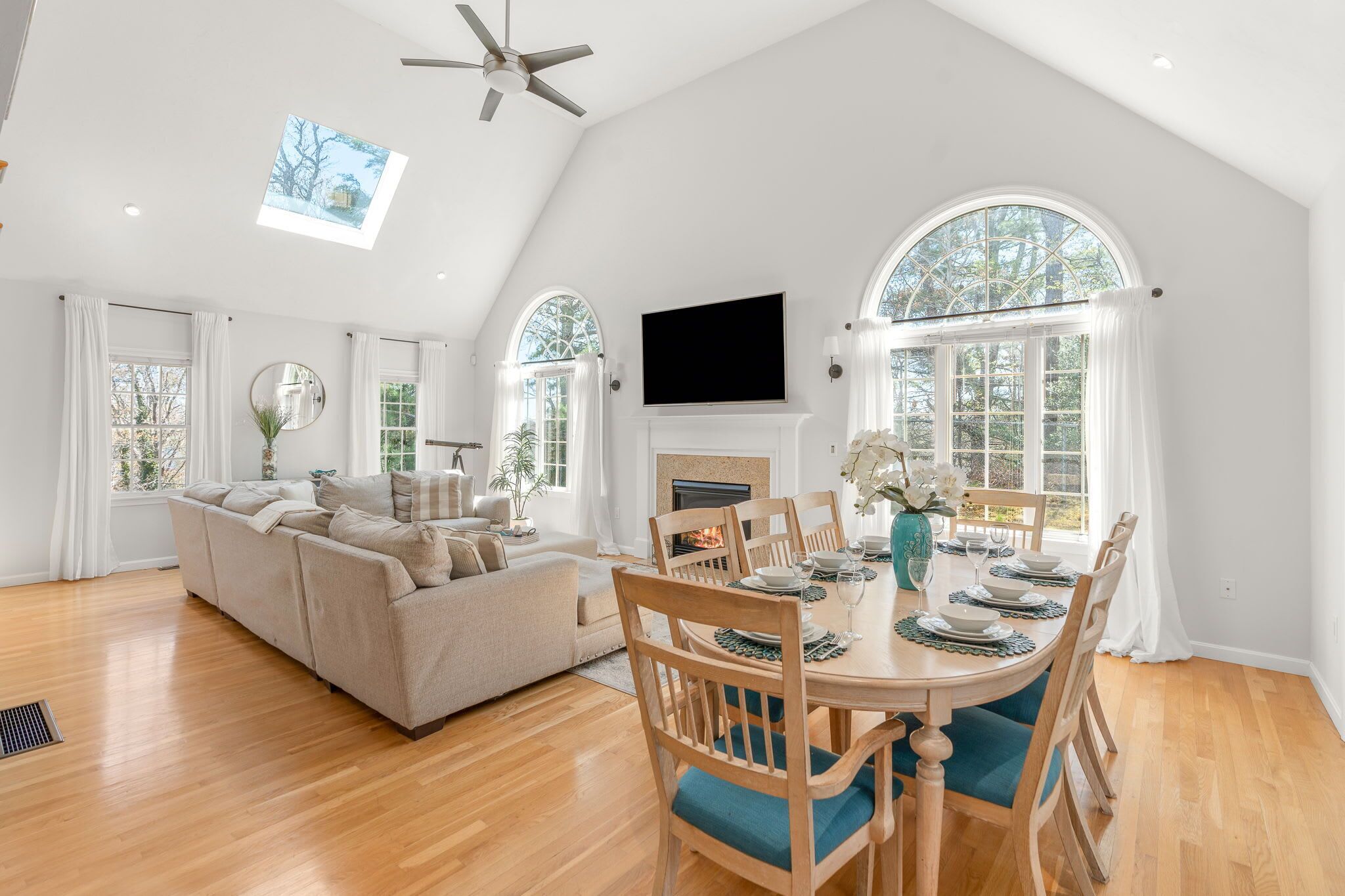 A spacious living room with a high ceiling, a fireplace, a dining area with a round table, and large arched windows.