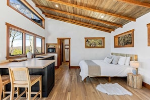 A bedroom with a bed, bedside table, and a view of trees through large windows. - Outdoor Movies w Hot Tub Fire Pit Mtn Views (Penrose)