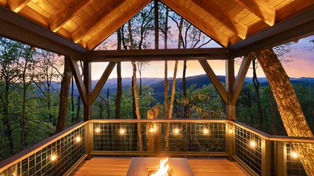 Una terraza de madera con una chimenea, rodeada de árboles y vistas a la montaña.