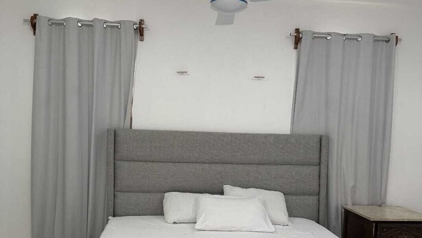 A bedroom with a bed, grey curtains, a ceiling fan, and a nightstand.