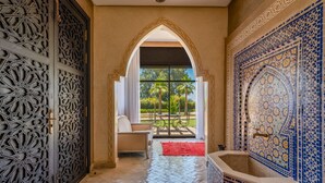 Un pasillo con una reja metálica decorativa, un área de estar con una mesa y sillas, vistas a palmeras y una pared de azulejos con diseños intrincados.