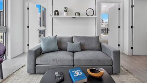 En moderne stue med en grå sofa, et sofabord med en bog og fjernbetjeninger, samt en hvid væg med en hylde over.