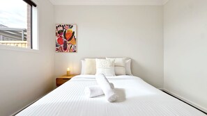 A hotel room with a bed, pillows, a nightstand with a lamp, and a colorful artwork on the wall. - Creekstone 4B Home by GoodLive (Tarneit)