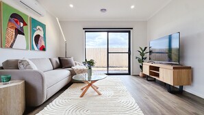 En moderne stue med sofa, sofabord i glas, tv på et træstel og et stort vindue.