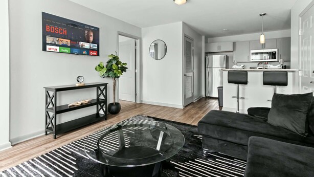 A modern living room with a black sofa, a glass coffee table, and a black and white striped rug. A flat-screen TV mounted on the wall displays various streaming services. - 3 mins from Arsenal/Bridge Street (Huntsville)
