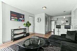 A modern living room with a black sofa, a glass coffee table, and a black and white striped rug. A flat-screen TV mounted on the wall displays various streaming services.