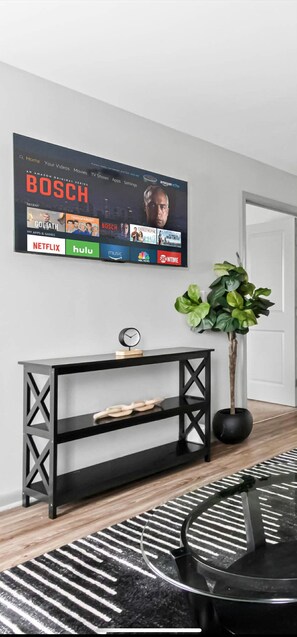 A living room with a mounted TV displaying a streaming service interface, a black console table, a potted plant, and a black and white rug. - 3 mins from Arsenal/Bridge Street (Huntsville)