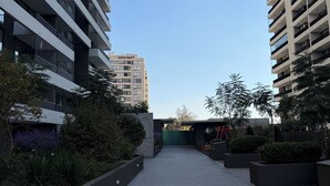 A modern urban building complex with multiple high-rise structures, a paved walkway, and landscaped areas with shrubs and trees. - Florida Metro Premium Studio + Parking + View (La Florida)