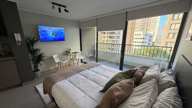 A modern bedroom with a large bed, a TV, a balcony, and a view of buildings. - Florida Metro Premium Studio + Parking + View (La Florida)