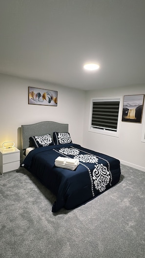 Un dormitorio con cama, una mesita de noche, una ventana con persianas y dos cuadros en la pared.