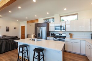 A modern kitchen with white cabinets, stainless steel appliances, a central island with stools, and a wooden ceiling. - Clearwater Summit: Panoramic Gorge Views & Modern Design (Hood River)