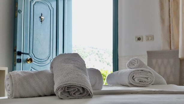 Een net opgemaakt bed met opgerolde witte handdoeken, een blauwe deur en uitzicht op groen.
