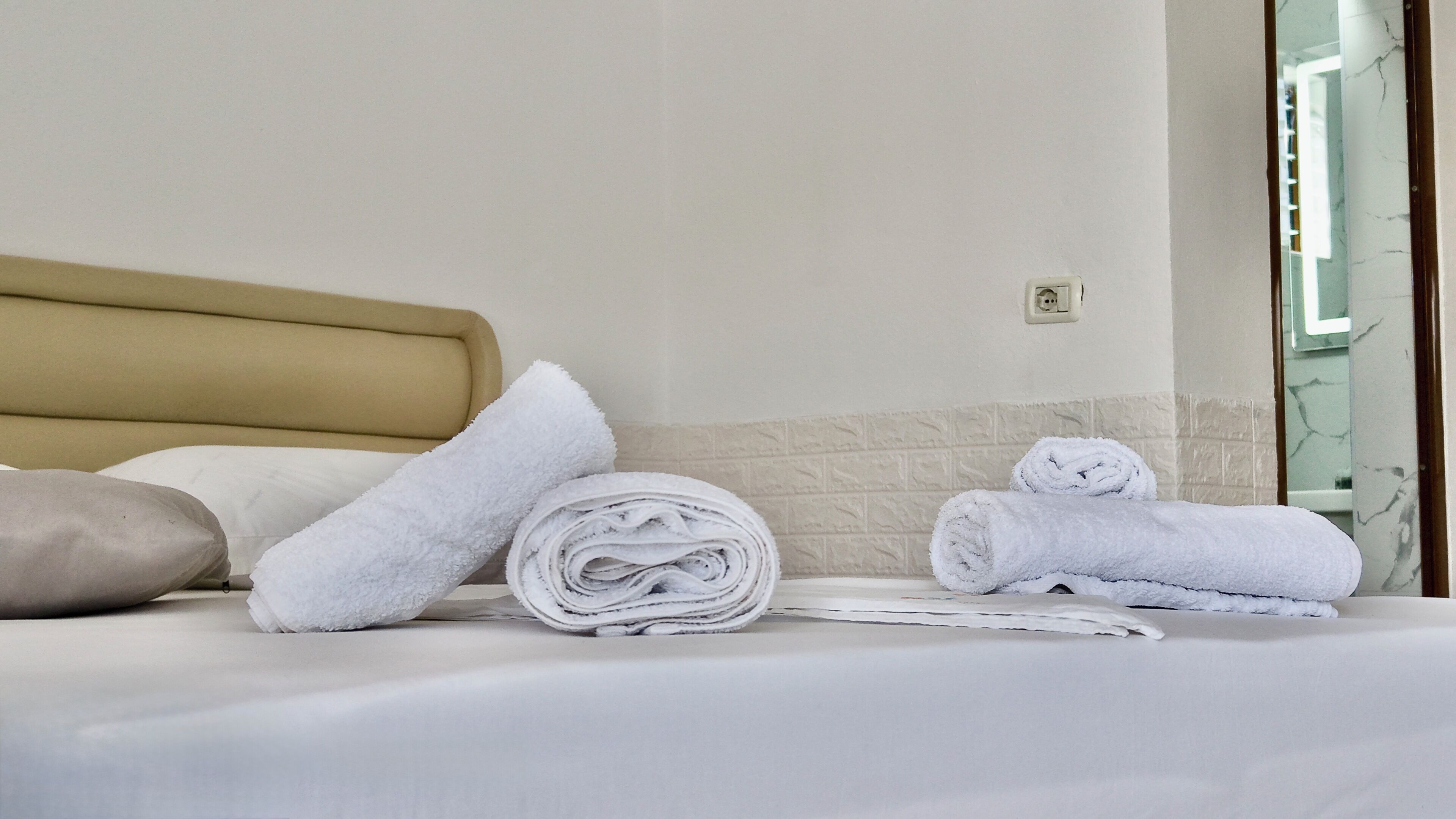 A neatly made bed with white linens, a beige headboard, and a rolled-up towel on the side.