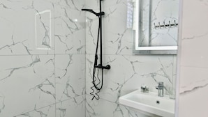 Een badkamer met witte marmeren tegels, een douche met een zwarte doucheslang, een wastafel met spiegel en een handdoekhouder.