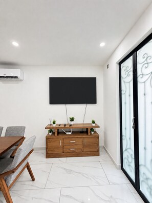 A modern living room with a flat-screen TV mounted on the wall, a wooden entertainment unit with plants, a dining table with chairs, and a sliding glass door with decorative metalwork. - REFUGIO HOUSE Tlaquepaque historic center a/c (Guadalajara)