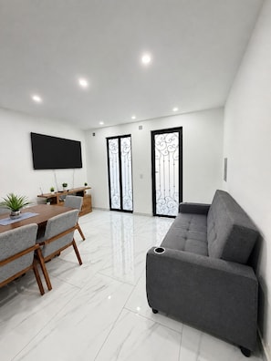 A modern living room with a grey sofa, a wooden dining table with chairs, a flat-screen TV mounted on the wall, and a sliding glass door with decorative metalwork. - REFUGIO HOUSE Tlaquepaque historic center a/c (Guadalajara)
