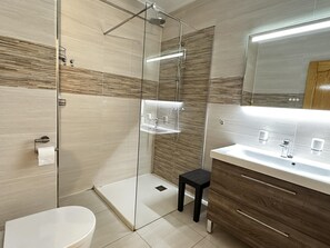 Et moderne badeværelse med en glasbrusekabine, toilet, håndvask med spejl og en lille skammel.
