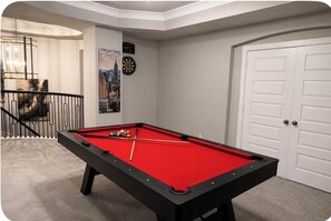 Ein Billardtisch mit roter Filzoberfläche, eine Dartscheibe und ein Stadtbild an der Wand.