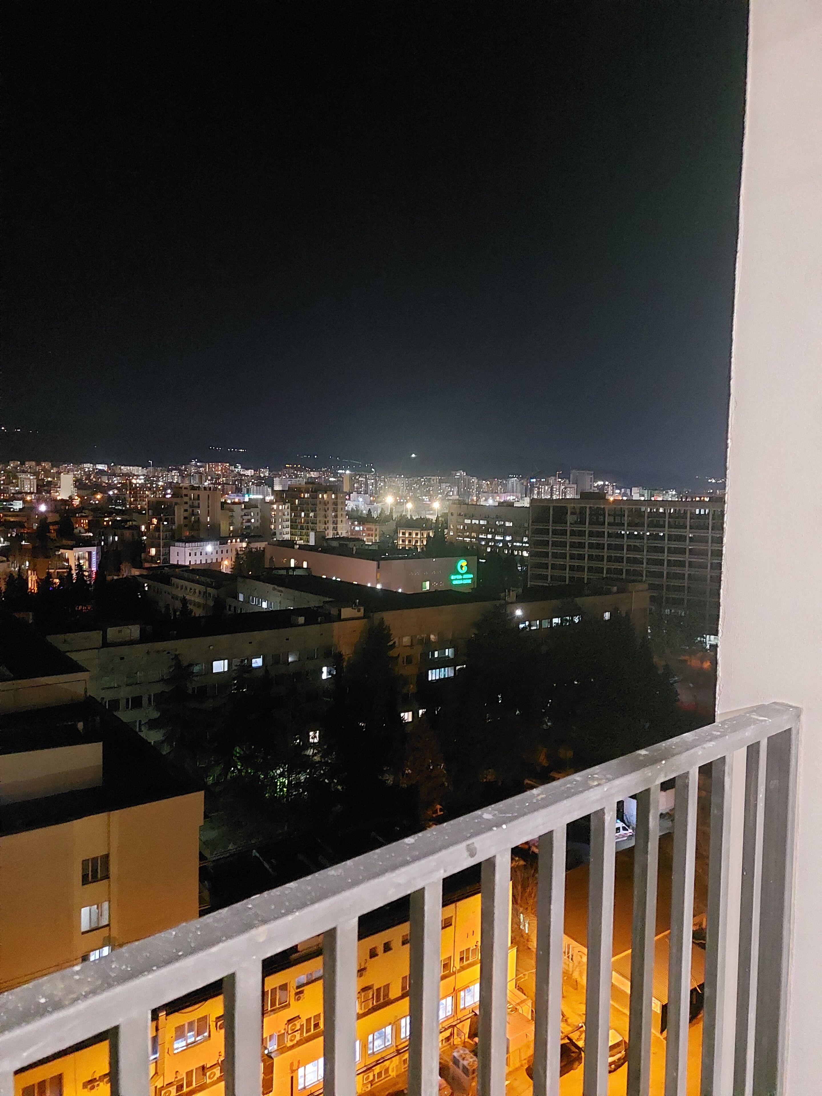 Une vue nocturne sur un paysage urbain, prise depuis un balcon, avec des bâtiments, une structure illuminée en vert et une rue bordée de voitures garées.