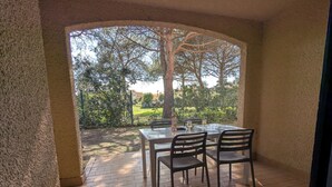 Eine überdachte Terrasse mit Tisch und Stühlen, einer Weinflasche und Blick auf einen Garten mit Bäumen und einem Zaun.