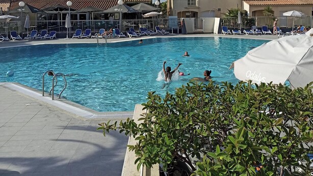 Ein Swimmingpool mit Liegestühlen und Sonnenschirmen, Palmen und Gebäude mit Ziegeldächern.