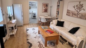 Ein Wohnzimmer mit Sofa, Couchtisch und Bücherregal. Es gibt einen Essbereich mit Tisch und Stühlen. Im Hintergrund ist eine Küche zu sehen.