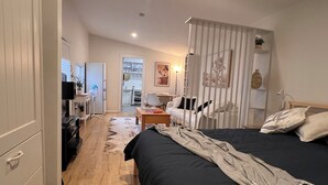 Ein Schlafzimmer mit einem Bett, einem Schreibtisch, einem Stuhl, einem Kleiderschrank und Blick zur Küche.