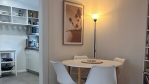 Ein Essbereich mit einem runden Tisch, weißen Stühlen, einem gerahmten Bild an der Wand, einer Stehlampe und einer Küche im Hintergrund.
