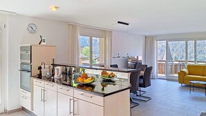 Eine moderne Küche mit Kochinsel, einem Essbereich mit gelbem Sofa und großen Fenstern mit Blick ins Freie.