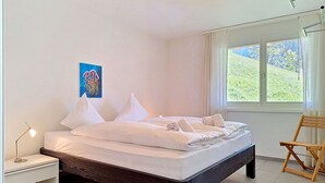 Ein Schlafzimmer mit Bett, Nachttisch, Sessel, Fenster mit Blick auf Grünflächen und einem Bild an der Wand.