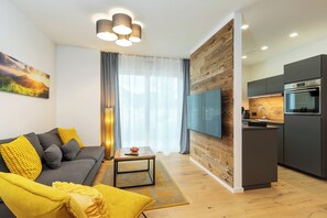 Ein modernes Wohnzimmer mit einem grauen Sofa, gelben Kissen, einem Couchtisch und einem Fernseher. Der Raum verfügt über Holzböden und ein großes Fenster mit Vorhängen.