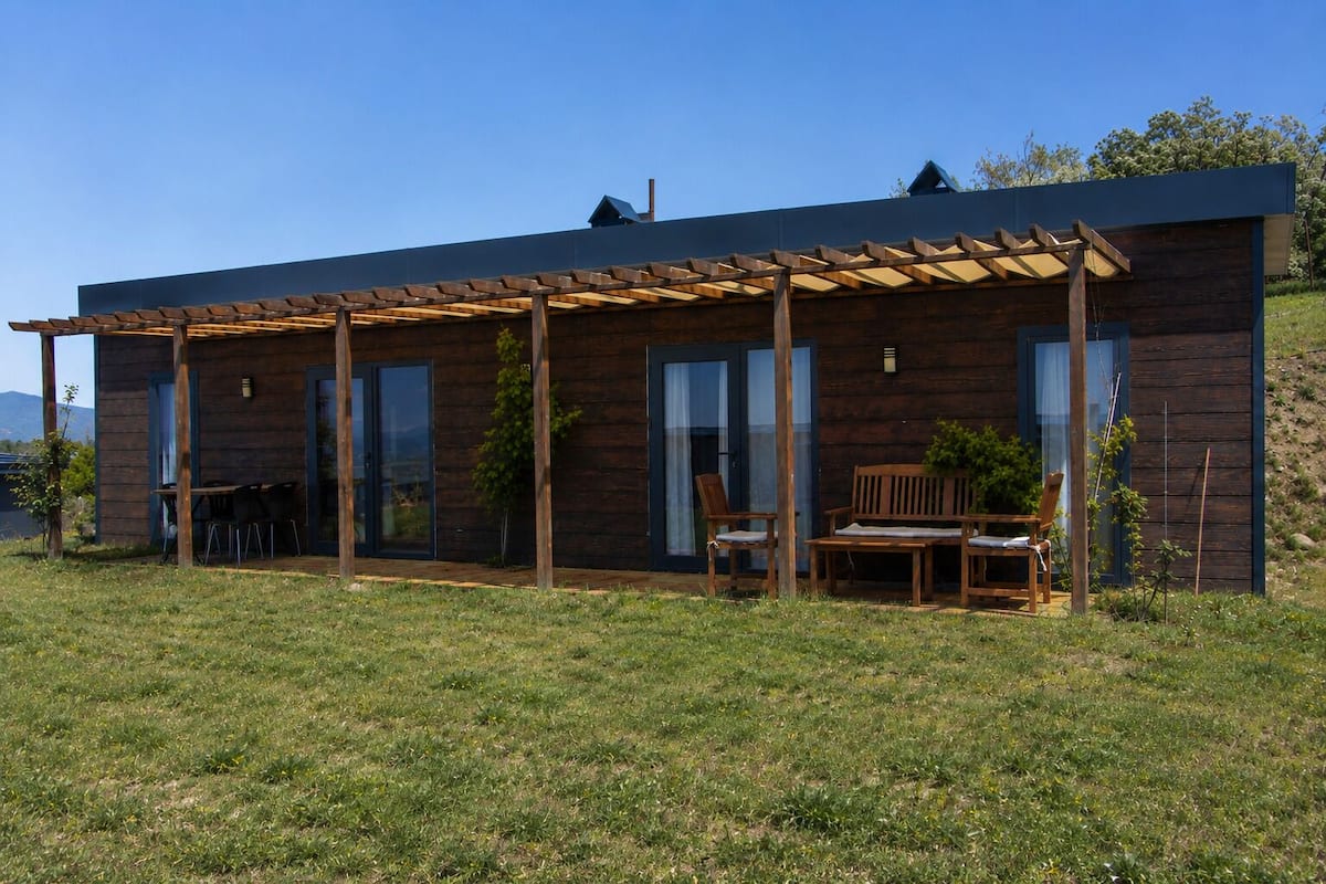Ein modernes Holzhaus mit einer überdachten Terrasse, Sitzgelegenheiten im Freien und Blick auf die umliegende Landschaft.
